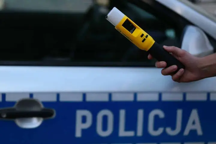 Dłoń trzymająca żółty alkomat na tle radiowozu policyjnego z napisem "POLICJA".