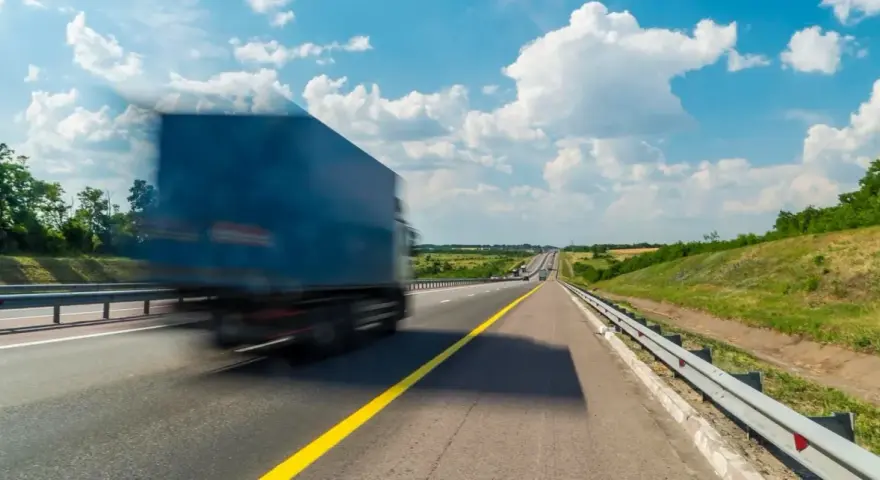 Niebieska ciężarówka w ruchu na autostradzie pod błękitnym niebem z chmurami.